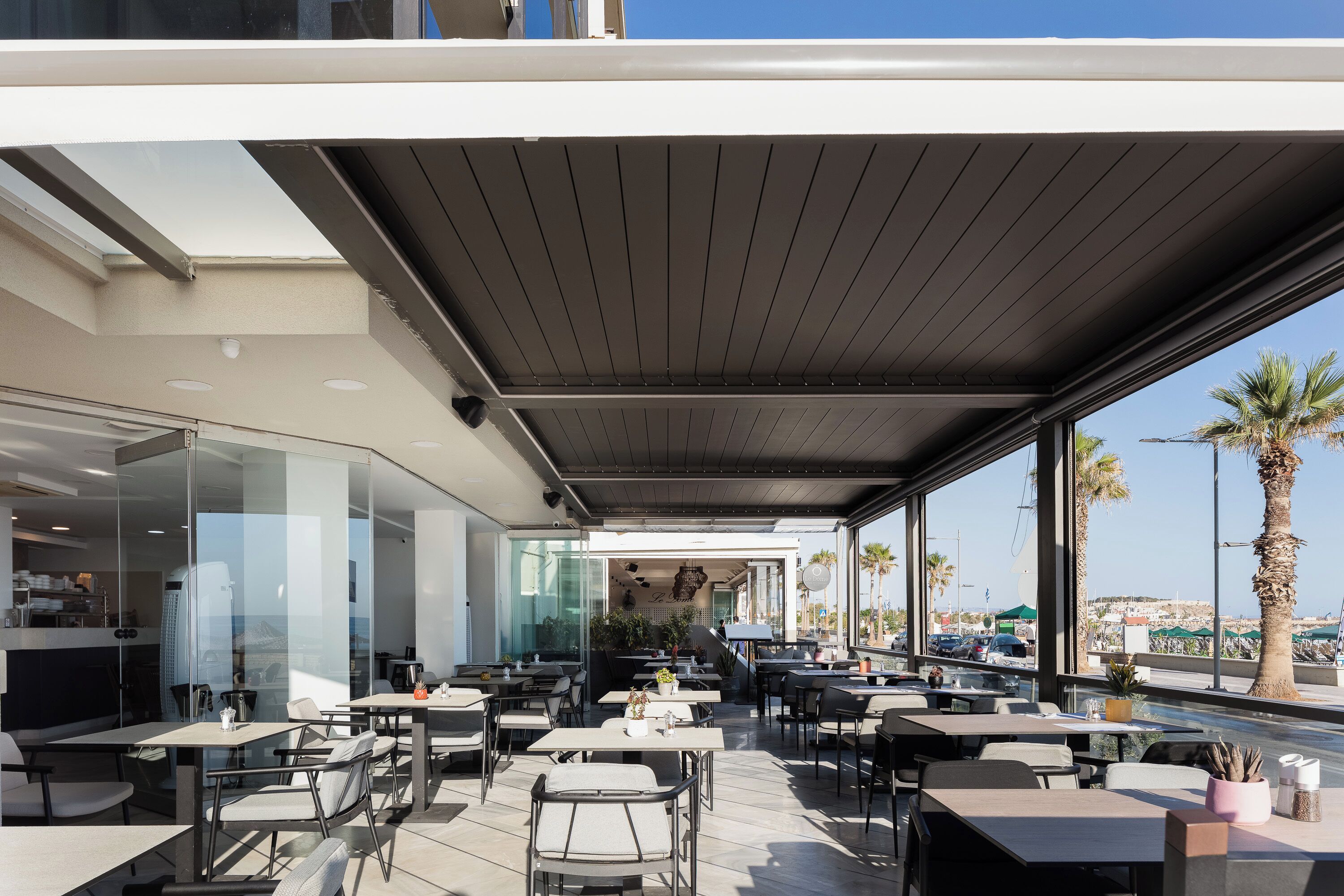 An outdoor dining area with tables and chairs, large windows, and a view of the sea.