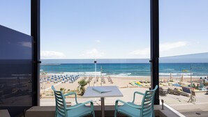 Een balkon met een tafel en stoelen, met uitzicht op een strand met parasols en ligbedden.