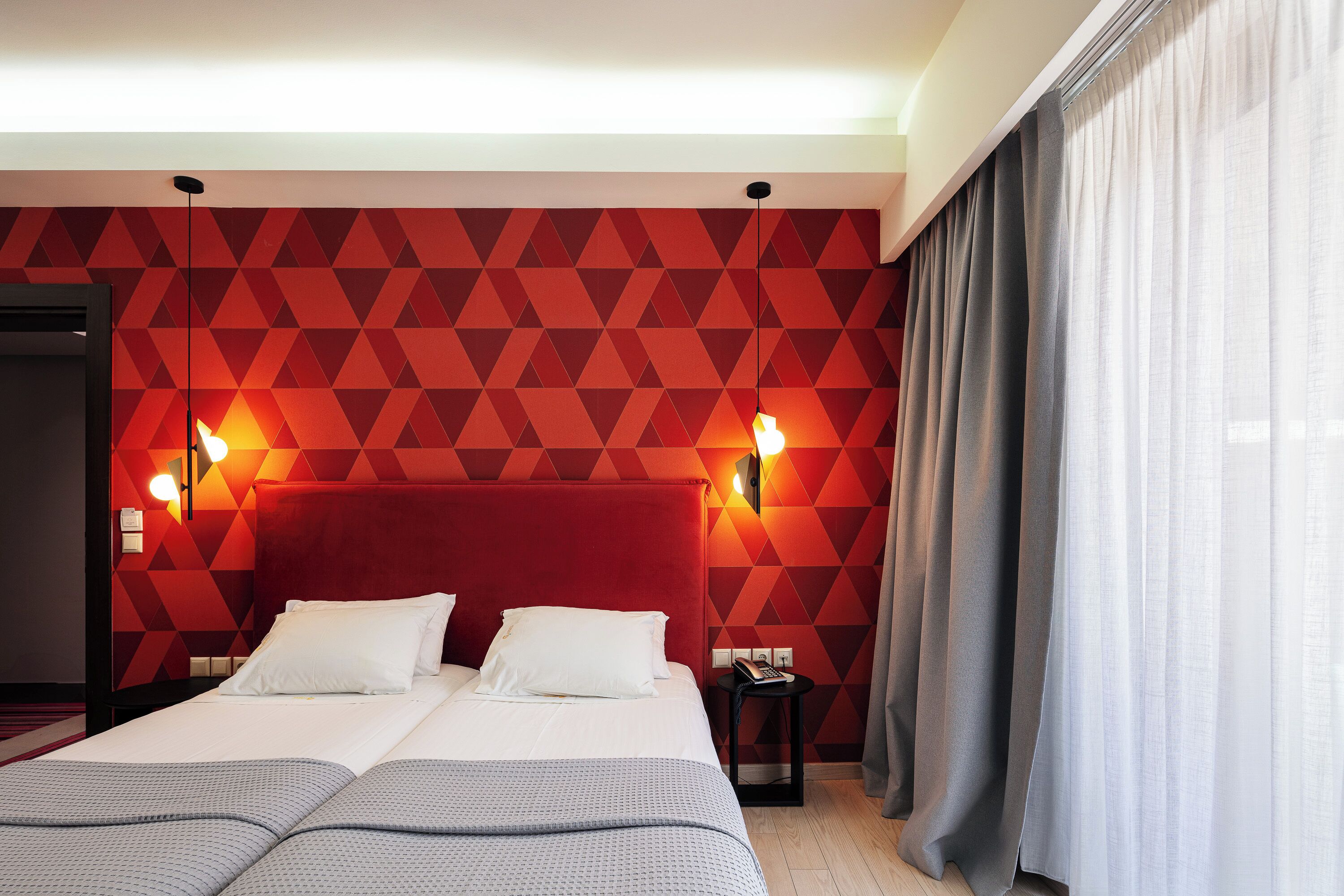 Habitación de hotel con un motivo geométrico rojo en la pared, una cama grande con dos almohadas, lámparas de noche y una mesita de noche con un libro.
