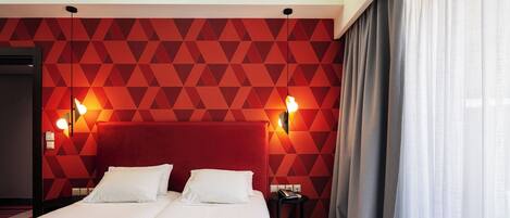 A hotel room with a red geometric wall pattern, a large bed with two pillows, bedside lamps, and a nightstand with a book.