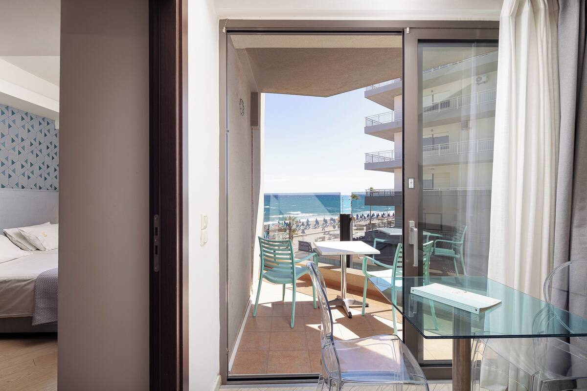 Un balcon avec une table et des chaises en verre, un lit avec une tête de lit à motifs et une vue sur la plage et l’océan.