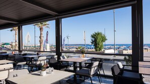Een terras met tafels en stoelen, met uitzicht op een strand met parasols en palmbomen.