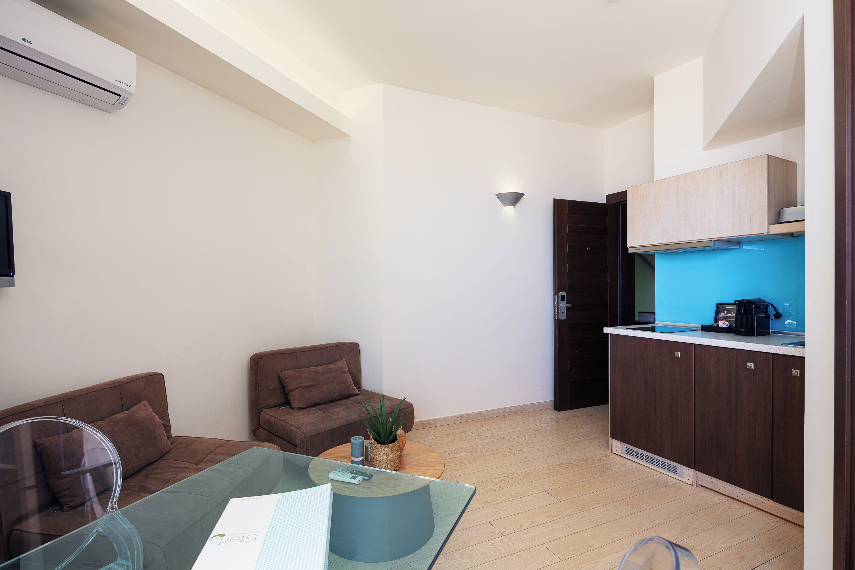 Una sala de estar moderna con una mesa de vidrio, dos sofás marrones, una mesita de centro con una planta y una cocina con armarios oscuros y un salpicadero azul.