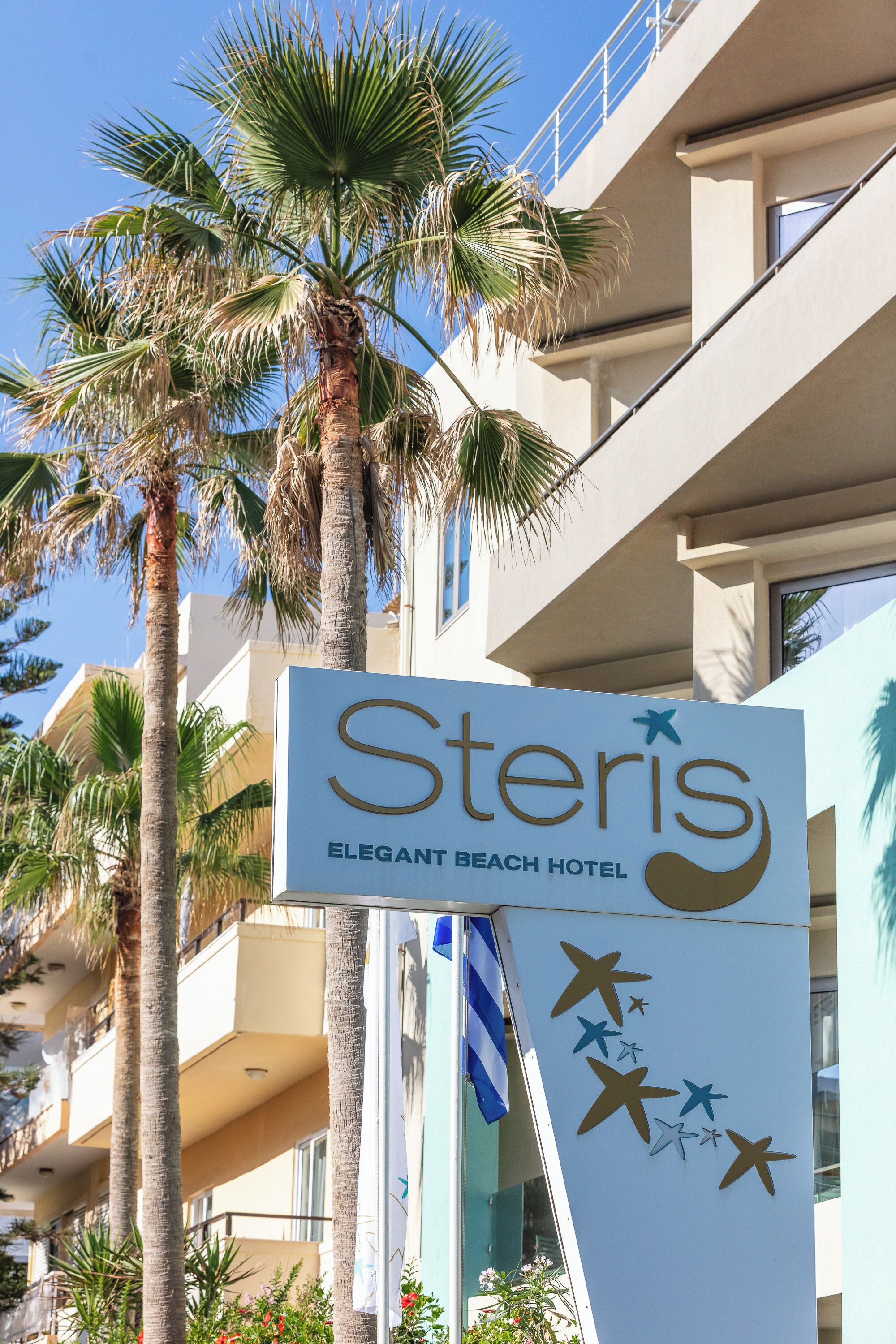 A sign for the Steris Elegant Beach Hotel with palm trees and a clear blue sky in the background.