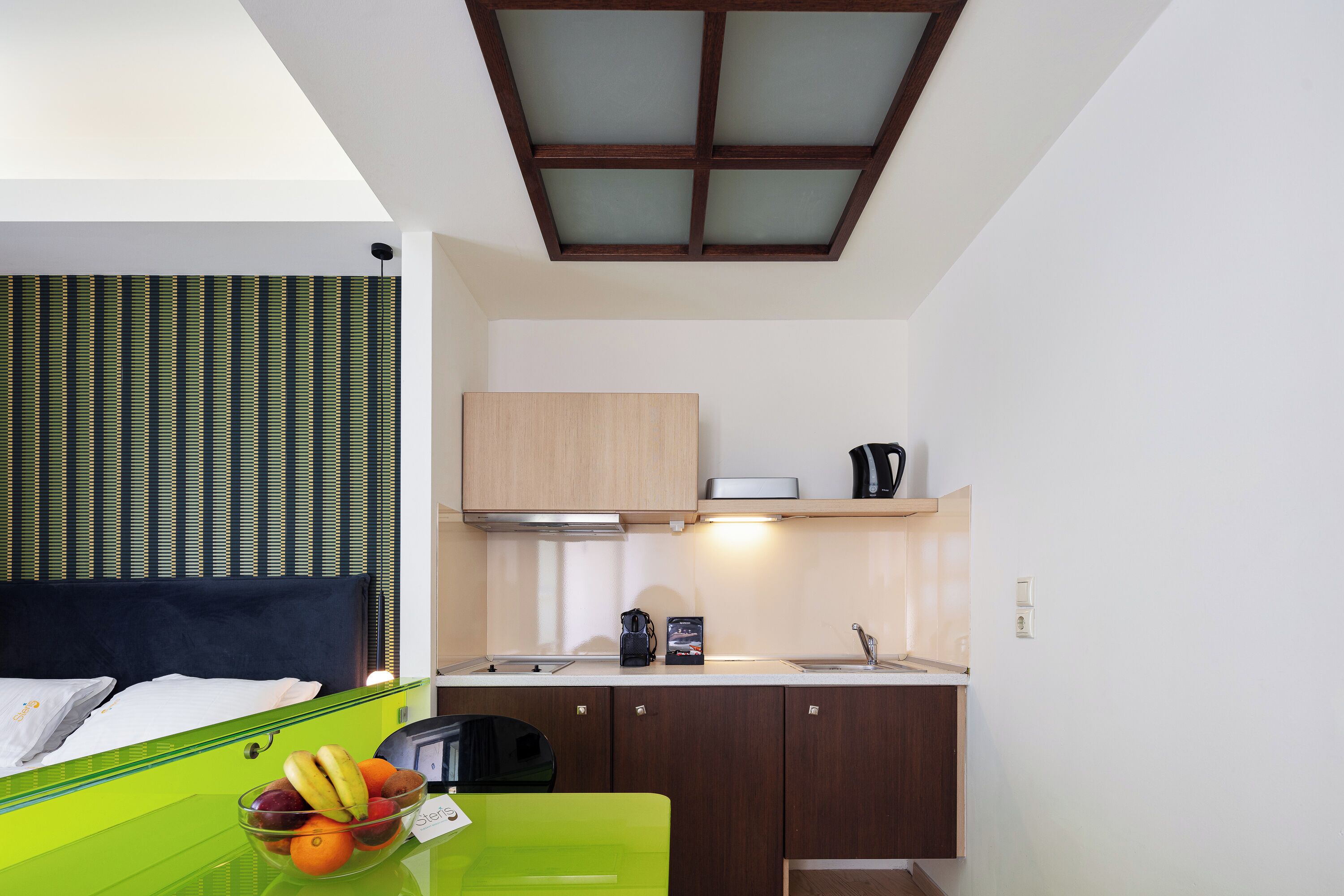 A compact hotel room with a kitchenette, a bed with a headboard, a green table with a fruit bowl, and a window with blinds.
