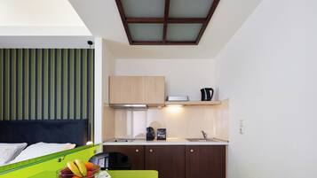 A compact hotel room with a kitchenette, a bed with a headboard, a green table with a fruit bowl, and a window with blinds.
