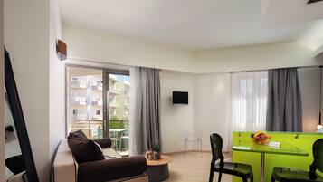 A modern living room with a sofa, a glass-top coffee table, a green desk with a fruit bowl, and a TV mounted on the wall.