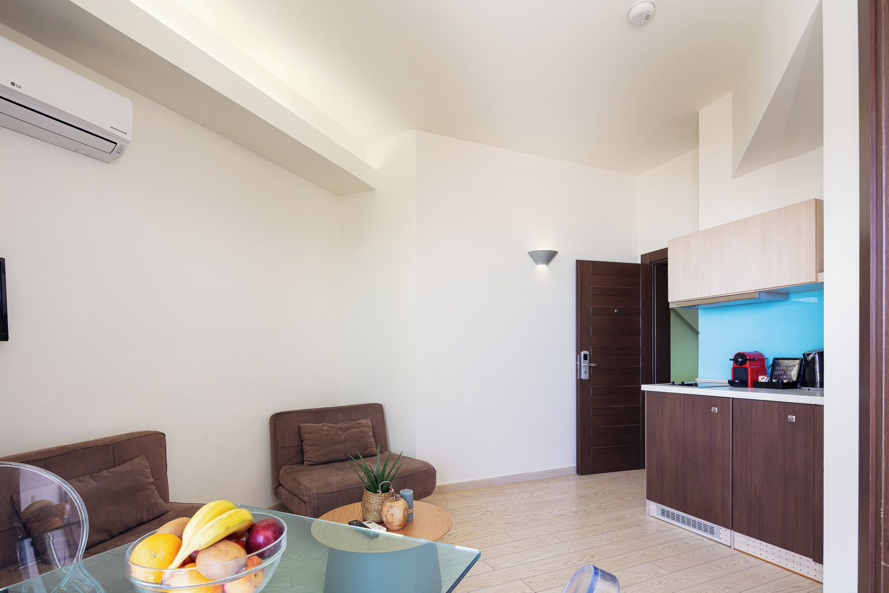 A modern living room with a glass coffee table, two brown sofas, a dining area with a bowl of fruit, and a kitchen with wooden cabinets and a blue backsplash.