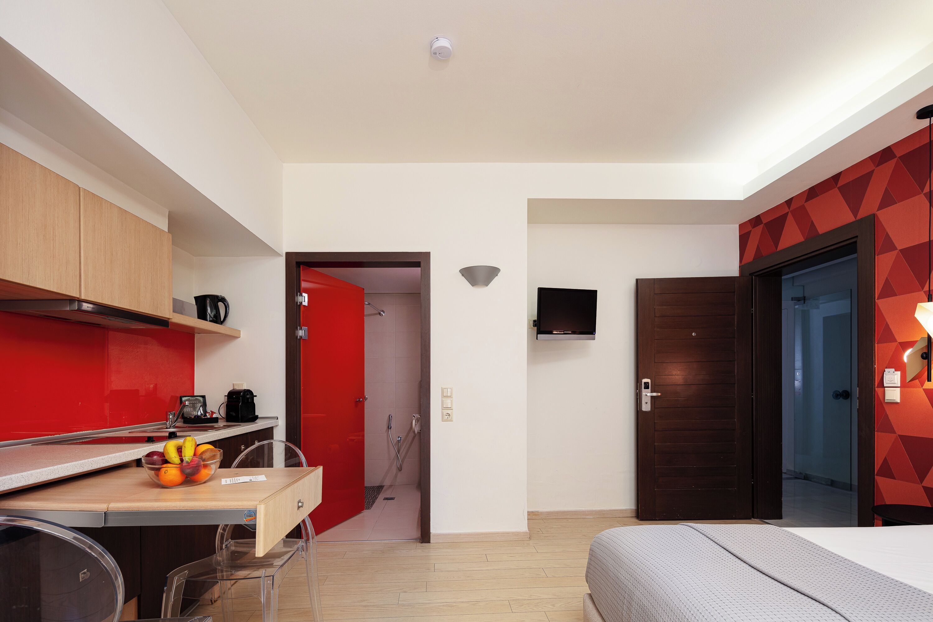 A compact hotel room with a kitchenette, a bed, a shower area, and a TV mounted on the wall.