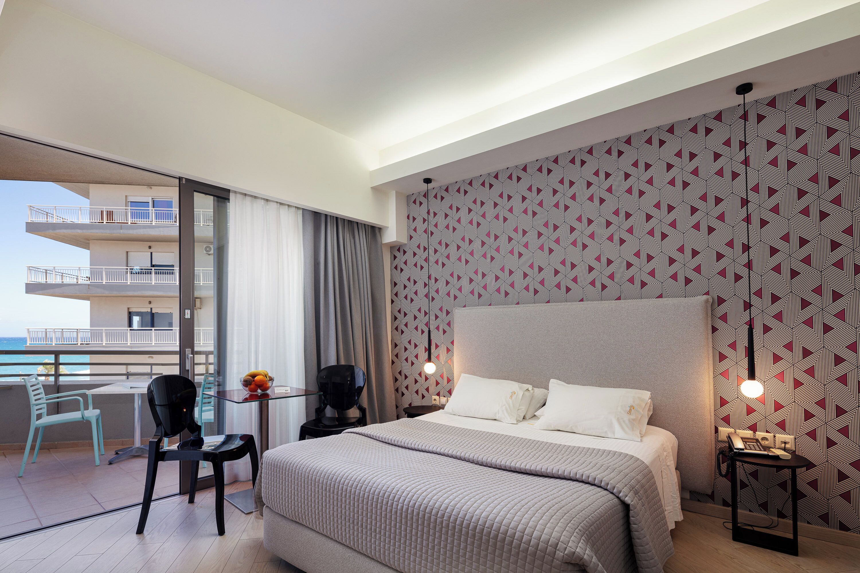 A modern hotel room with a large bed, a balcony with a view, a small table with a chair, and a patterned wall.