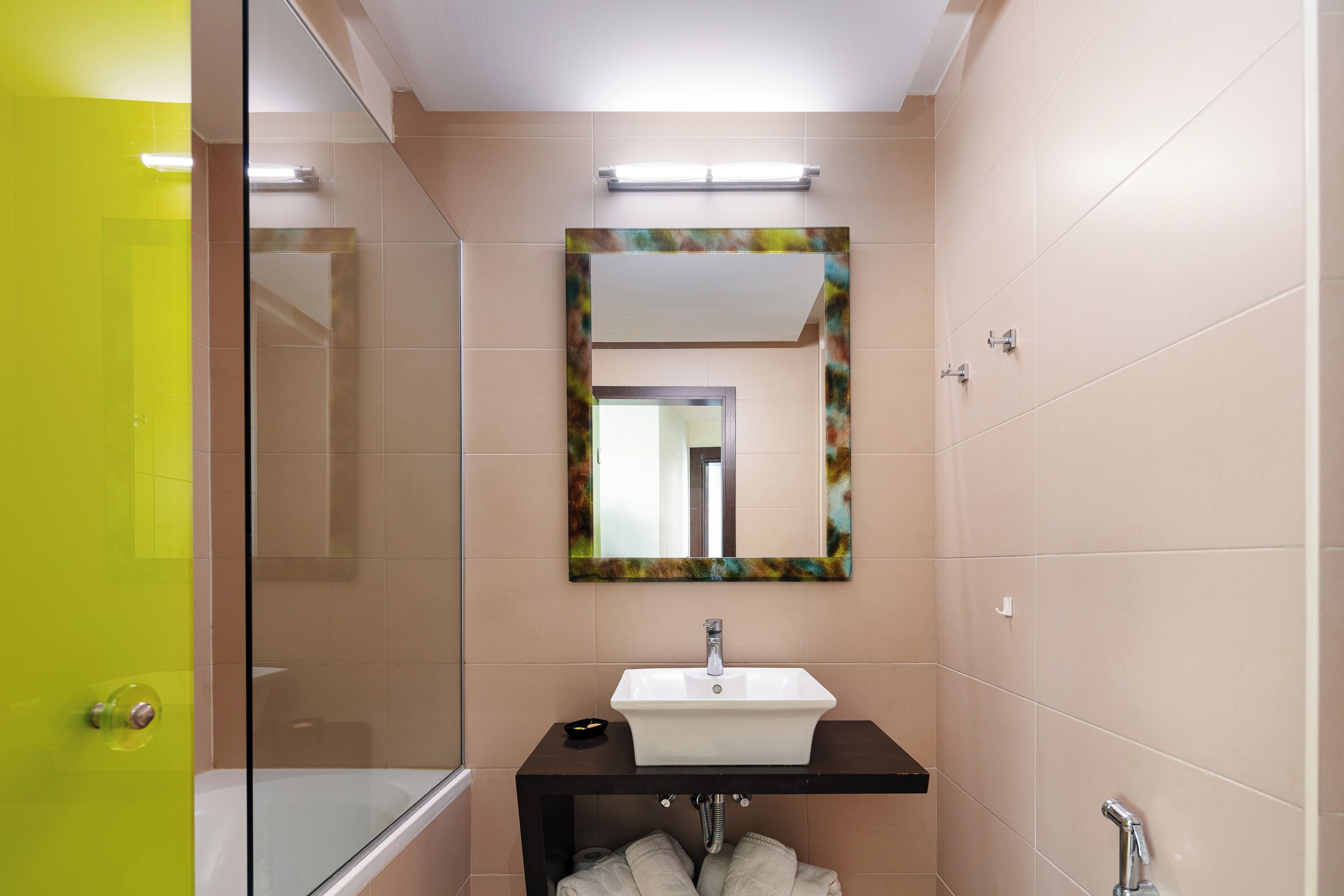 A bathroom with a glass shower enclosure, a white sink with a black countertop, and a mirror with a colorful frame.