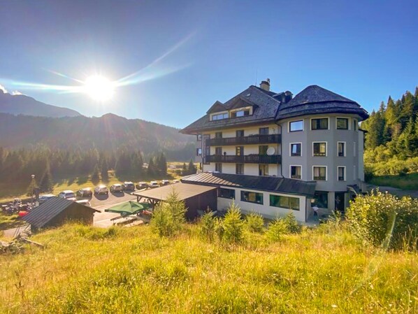 A multi-story hotel with a curved roof, located in a mountainous area with a parking lot and a small building in the foreground. - Al Gallo Forcello 1530 (Pontebba)