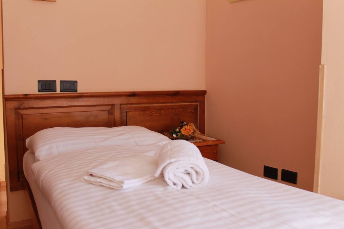 A neatly made bed with a white blanket and a rolled towel, a wooden headboard, and a small bedside table with a flower vase.