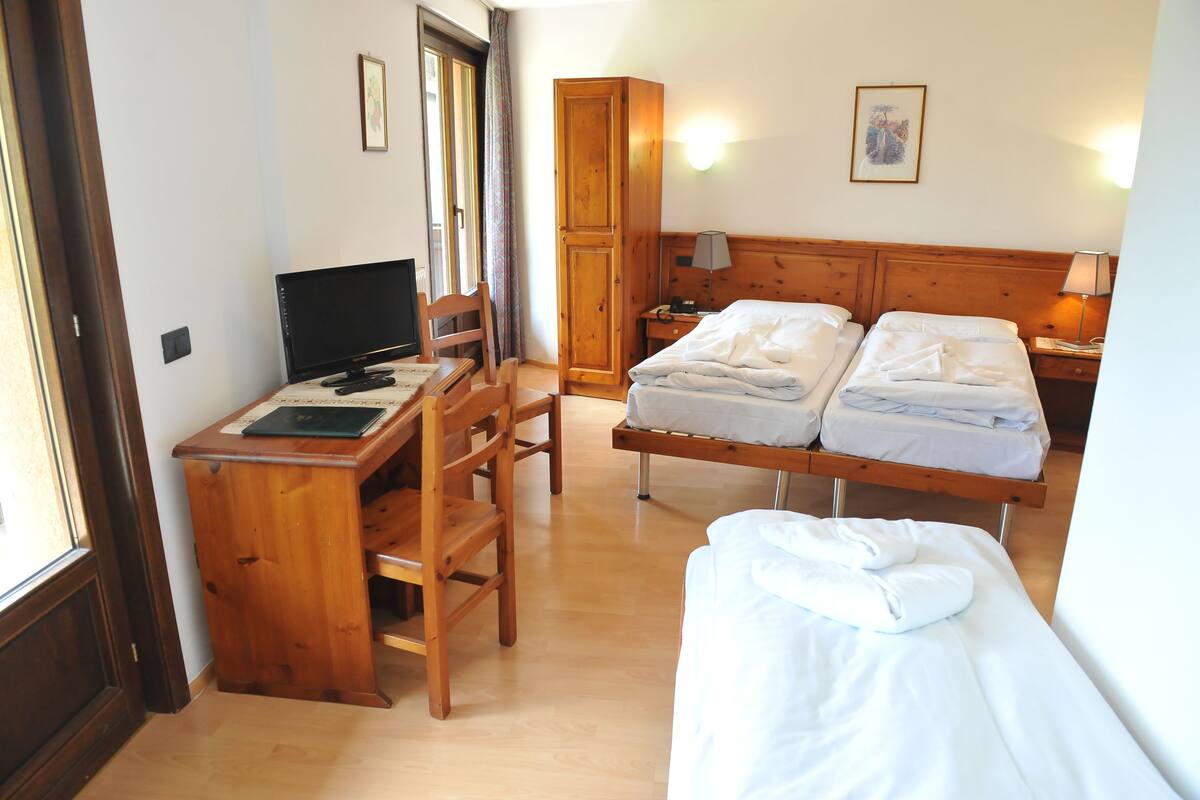 A hotel room with a wooden desk, two beds, a wardrobe, and a television.