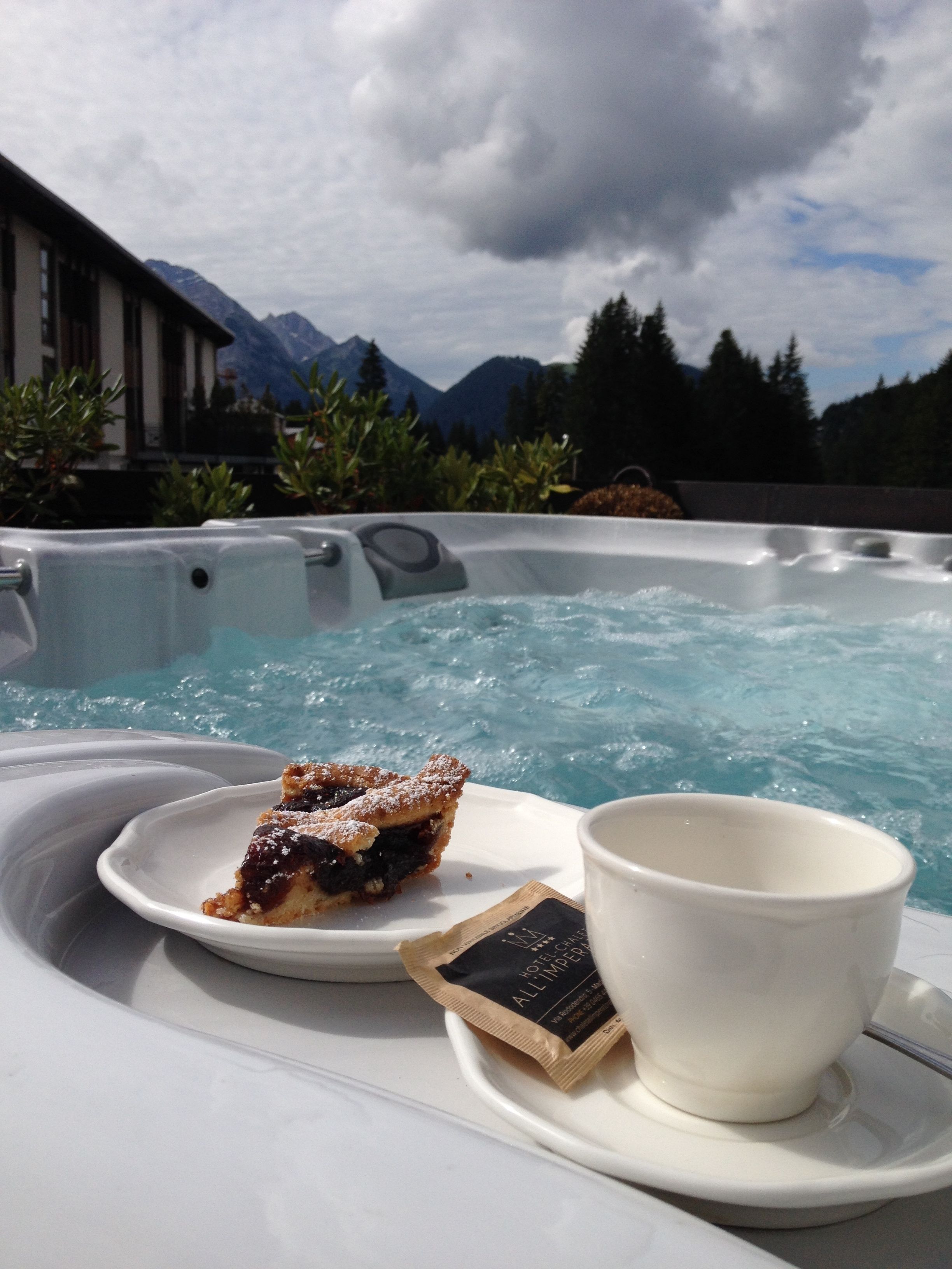 A hot tub with a slice of pie and a cup on a tray, set against a backdrop of mountains and trees.