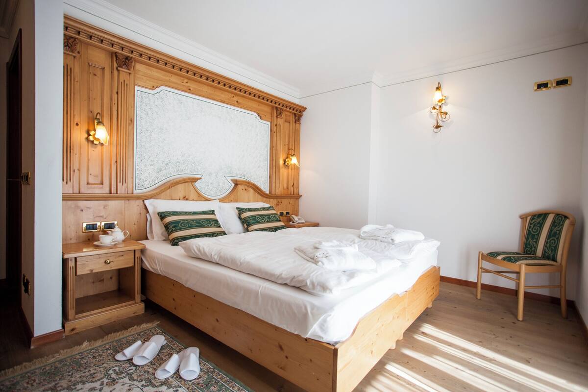A wooden bed with white linens, a nightstand with a lamp, and a chair in a room with wooden floors and white walls.