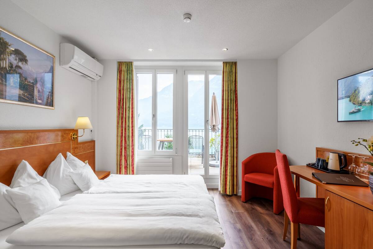 A hotel room with a large bed, a desk with a chair, a TV, and a balcony with curtains.