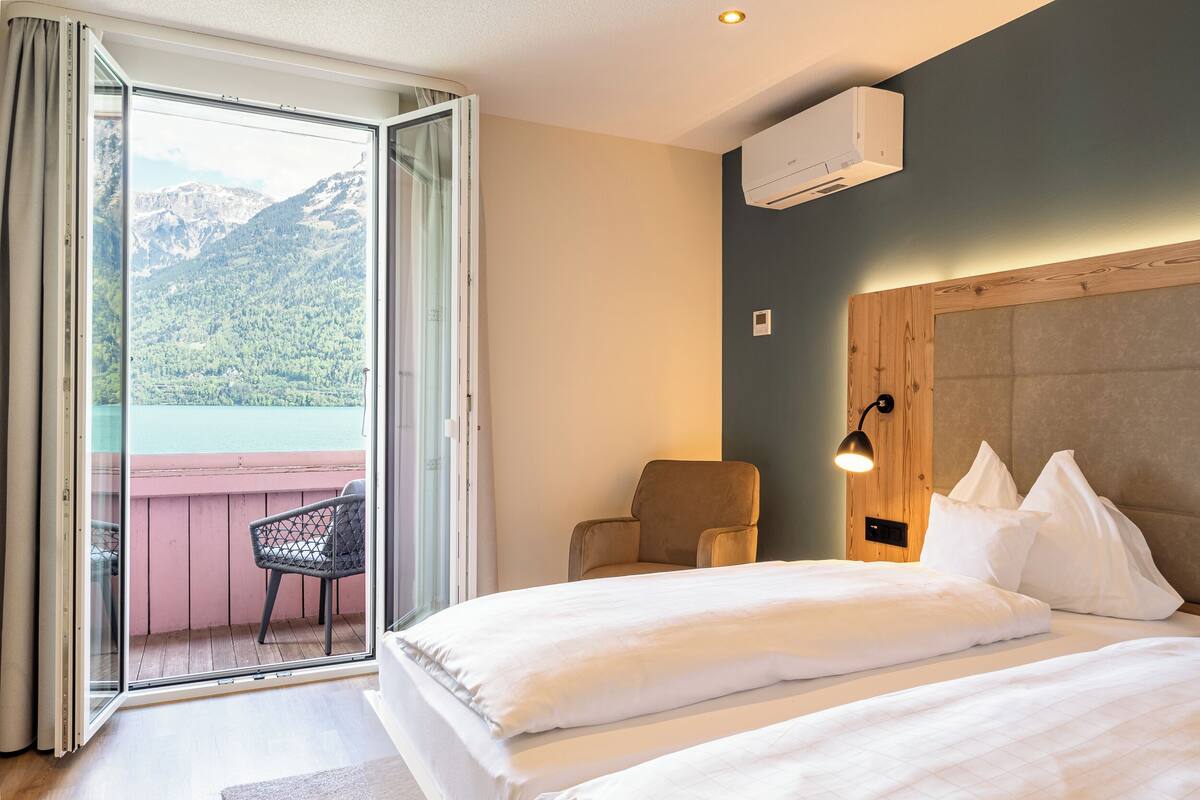 A hotel room with a bed, a chair, a wooden headboard, and a view of mountains through a sliding glass door.