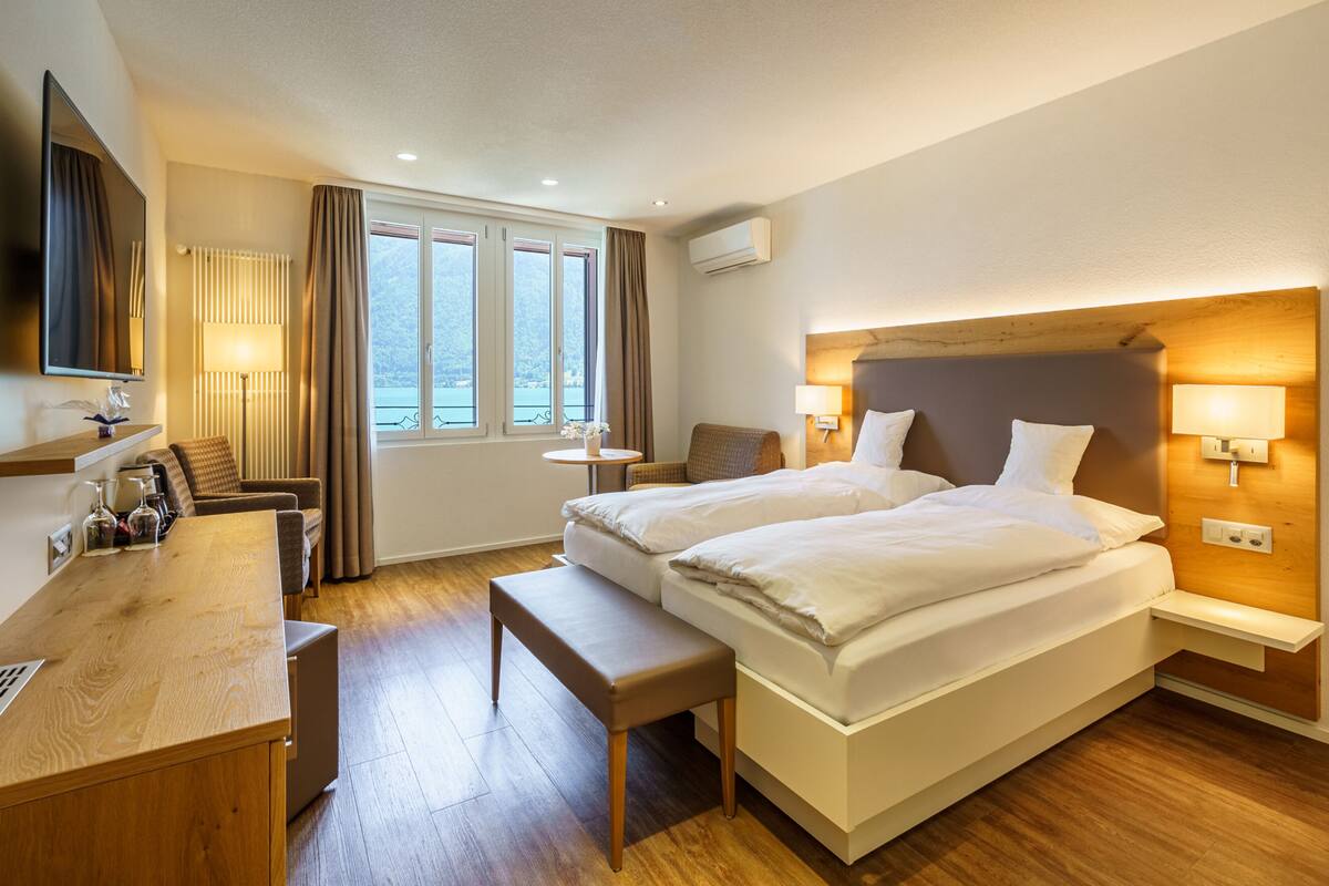 A hotel room with two beds, a desk, a chair, a small table, and a view of the outdoors through large windows.