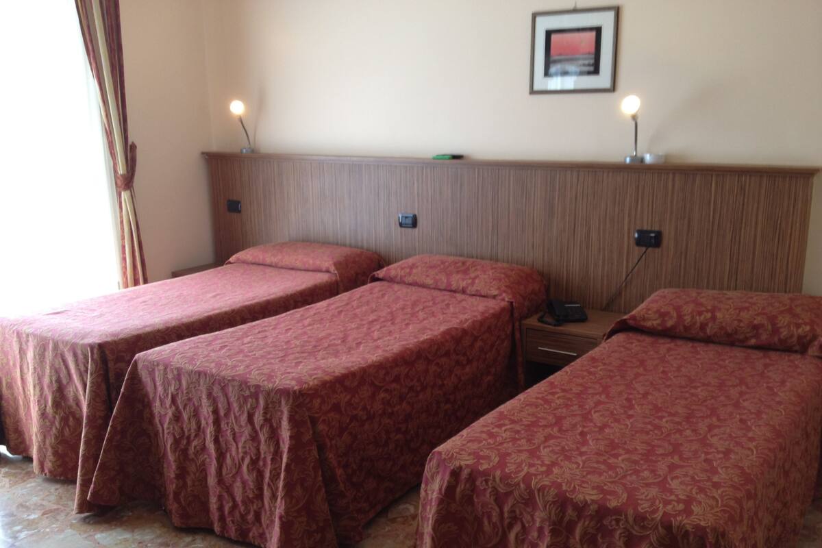 A hotel room with three single beds, a wooden headboard, bedside lamps, a framed picture on the wall, and a telephone on the nightstand.