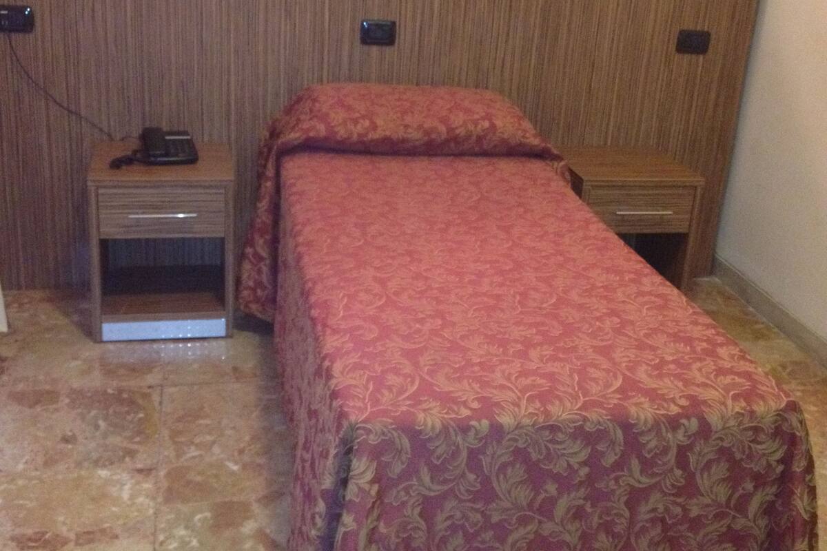 A single bed with a patterned bedspread, a nightstand with a phone, and a wall-mounted light fixture.