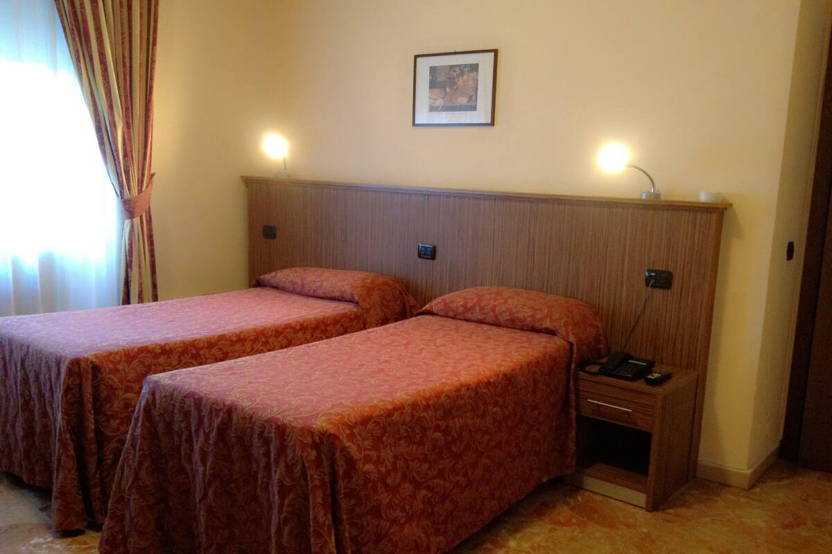 A hotel room with two single beds, a nightstand, and a wall-mounted lamp.