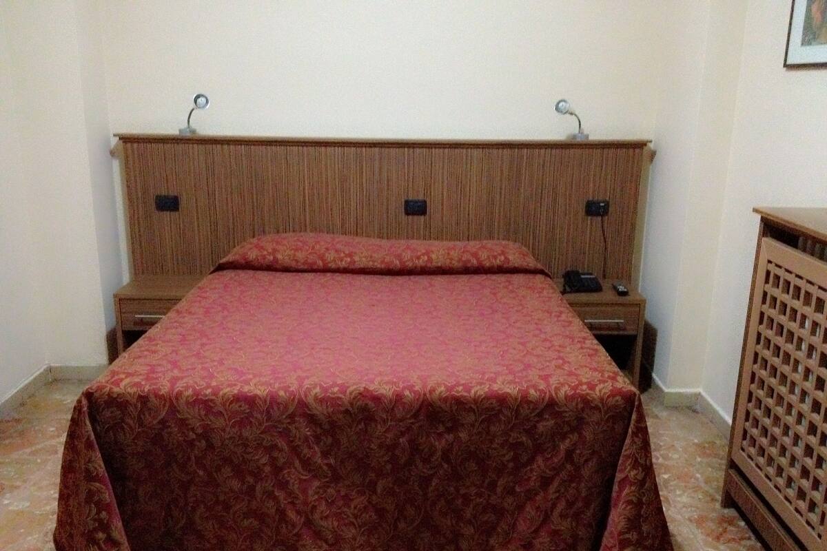 A bedroom with a wooden headboard, a red bedspread, two bedside tables, and a wooden cabinet.
