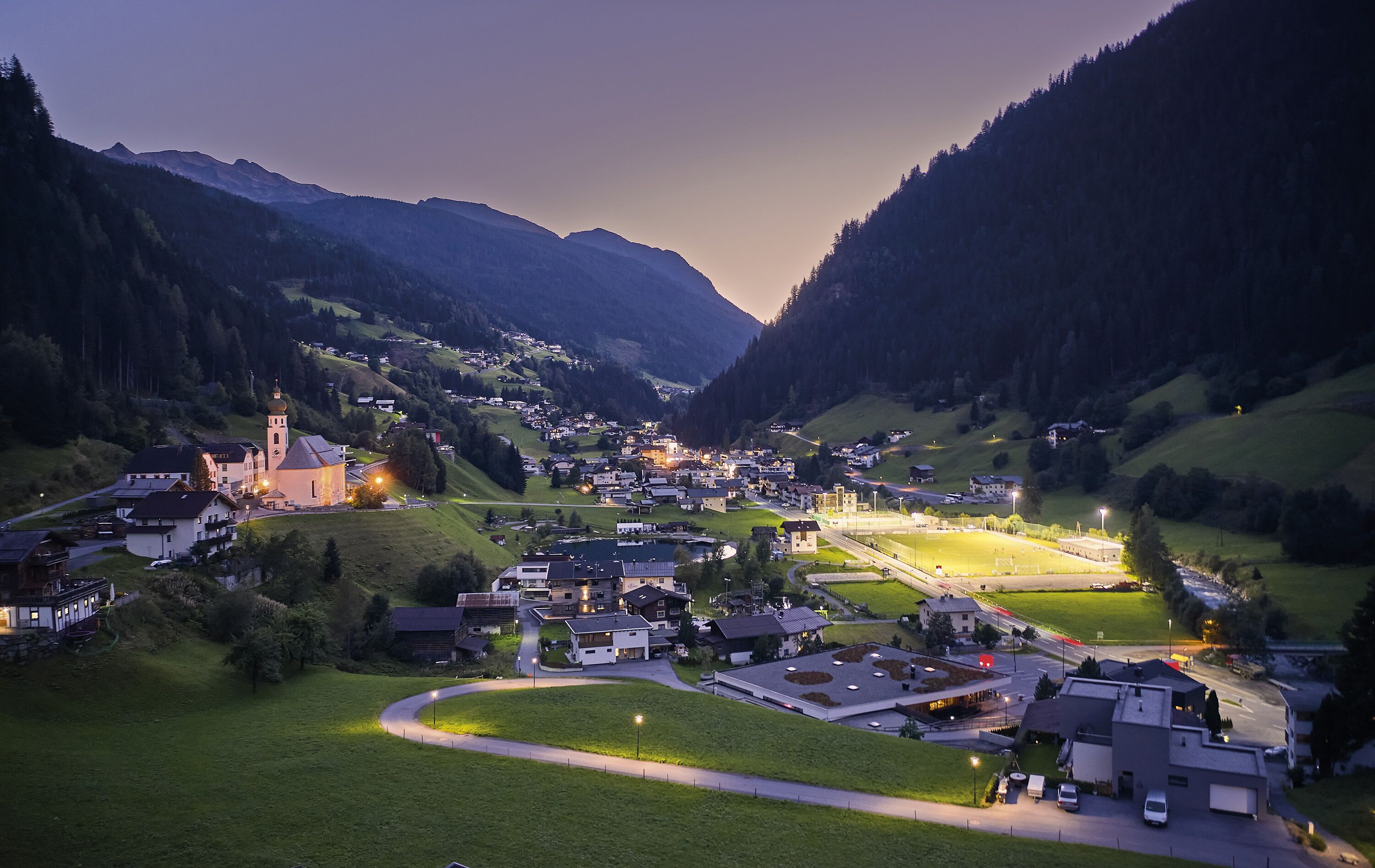A town nestled in a valley with a prominent church, surrounded by mountains and lit by streetlights.