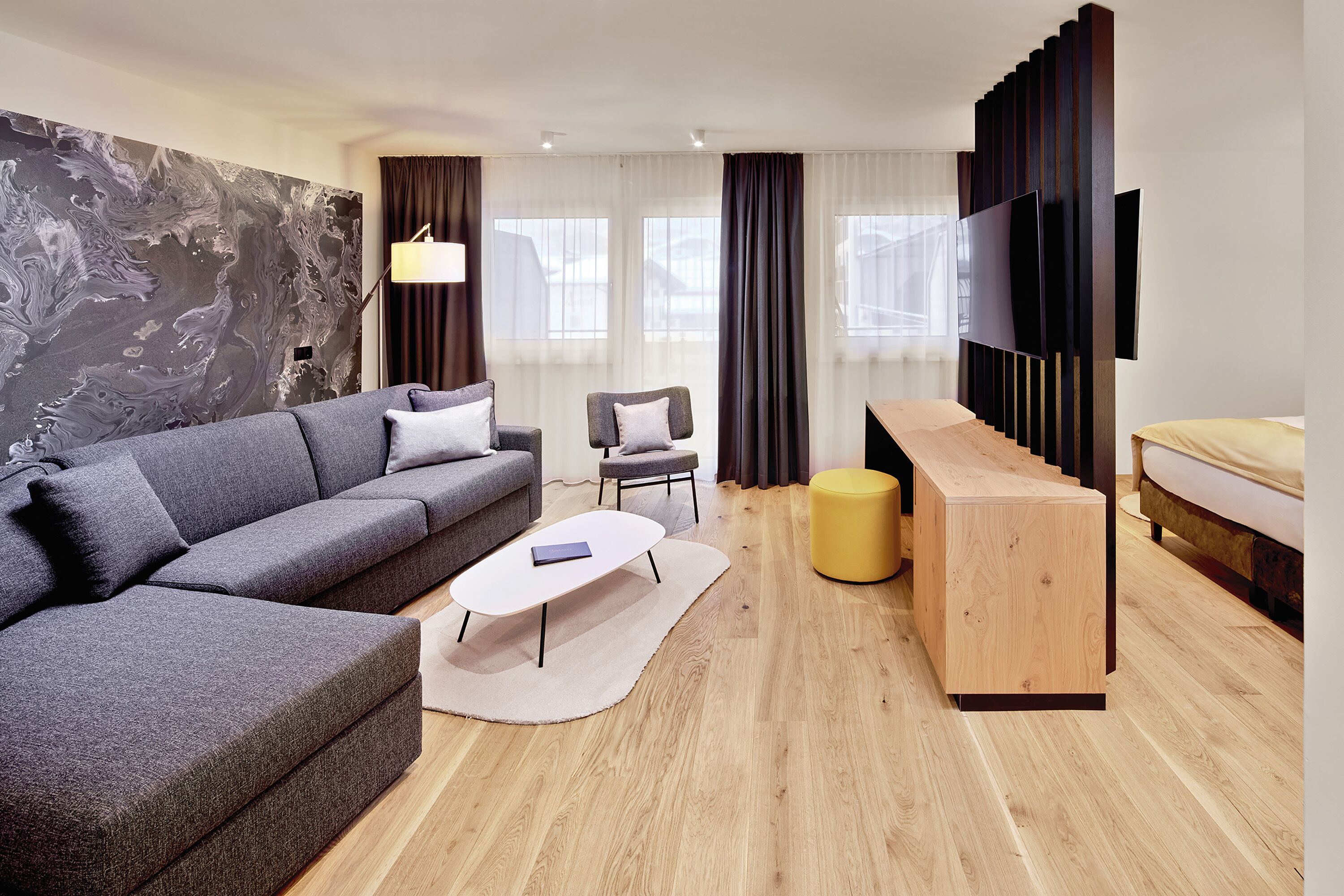 Una sala moderna con un sofá gris, una mesita de centro pequeña, una mesita auxiliar de madera, un reposapiés amarillo y un televisor de pantalla plana montado en la pared.