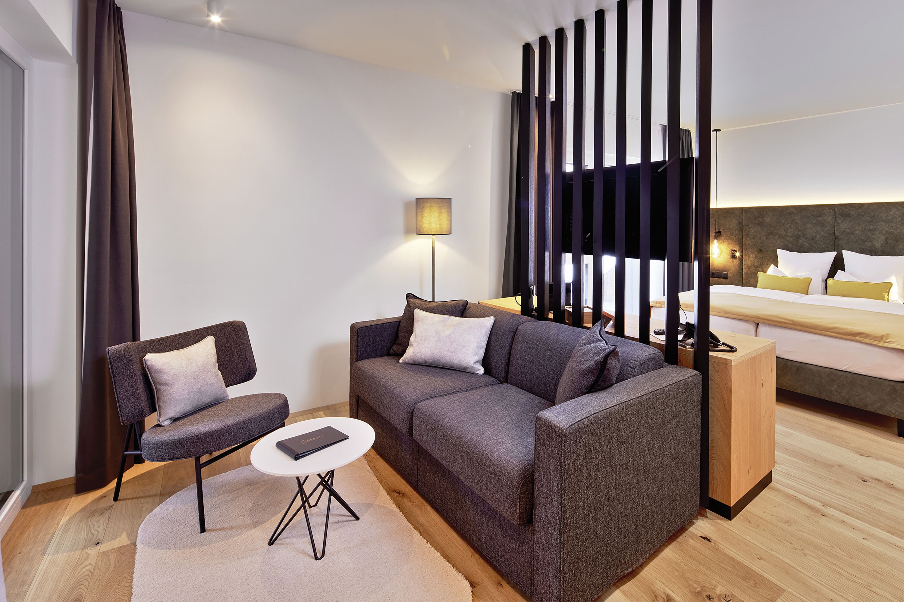 A modern hotel room with a grey sofa, a small round table, a wooden side table, a bed with white and yellow bedding, and a large window with vertical blinds.