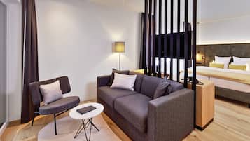 A modern hotel room with a grey sofa, a small round table, a wooden side table, a bed with white and yellow bedding, and a large window with vertical blinds.