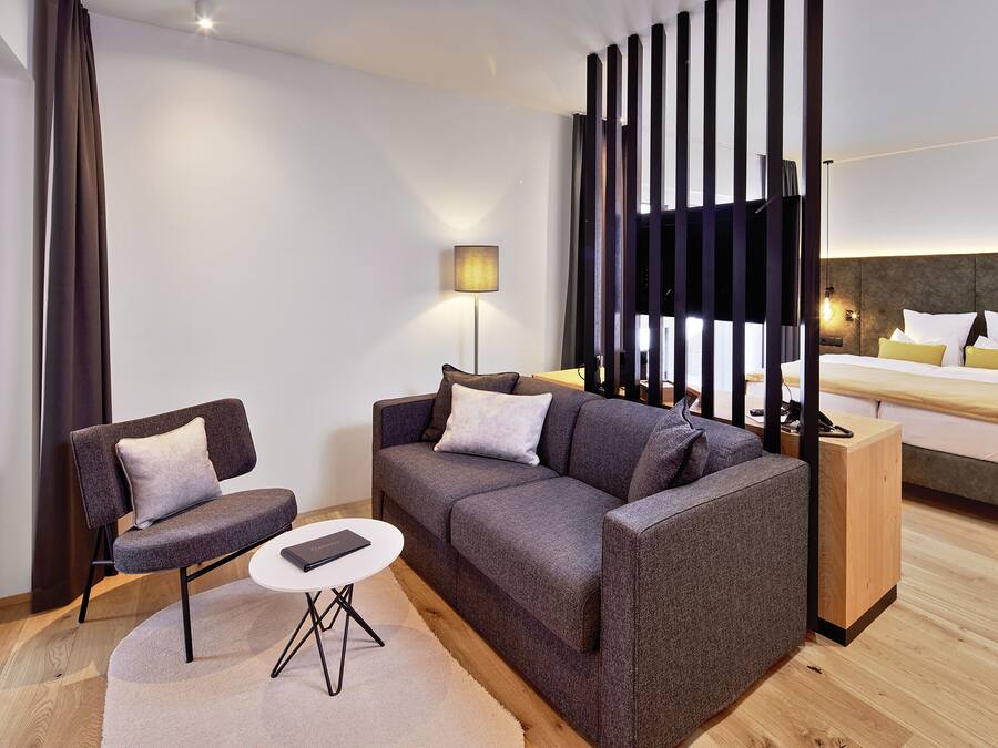 Una habitación de hotel moderna con un sofá gris, una mesa redonda pequeña, una mesita de noche de madera, una cama con ropa de cama blanca y amarilla, y un ventanal con persianas verticales.