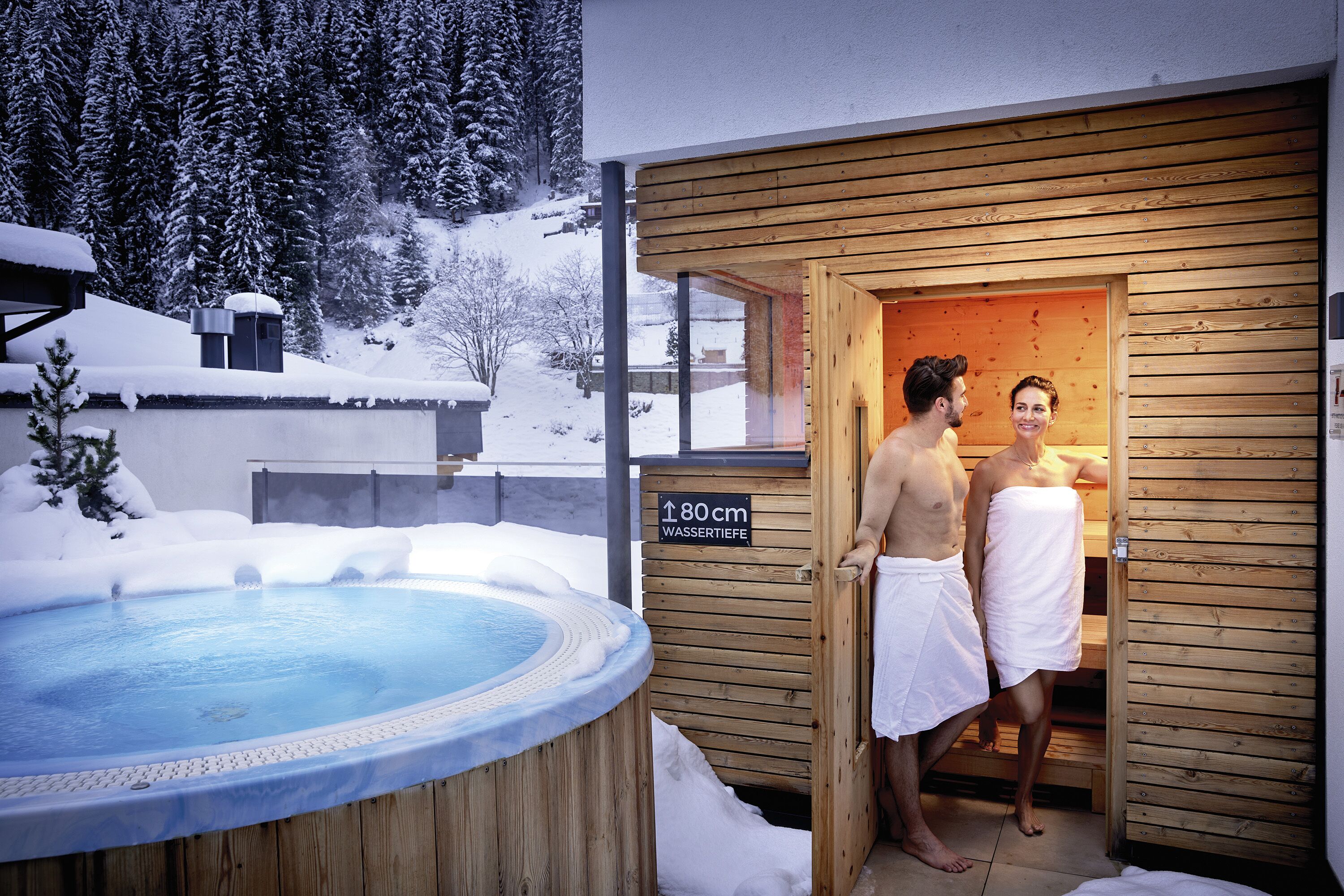 A wooden sauna with a sign indicating a height of 80 cm, a hot tub, and a snowy outdoor setting.