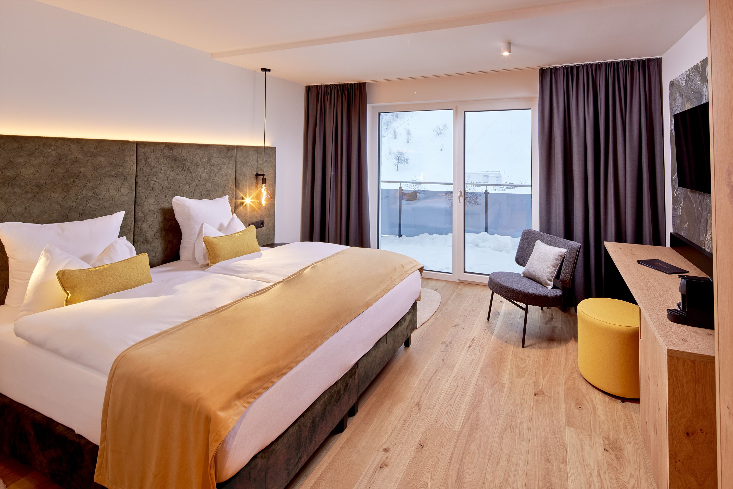A modern hotel room with a large bed, a desk, a chair, and a view of the outdoors.