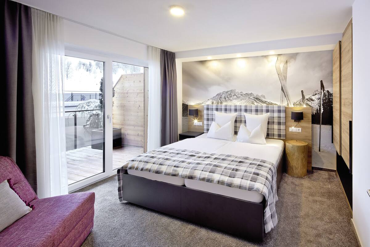 A modern bedroom with a large bed, a sofa, a wooden side table, and a view of a snowy landscape through a sliding door.