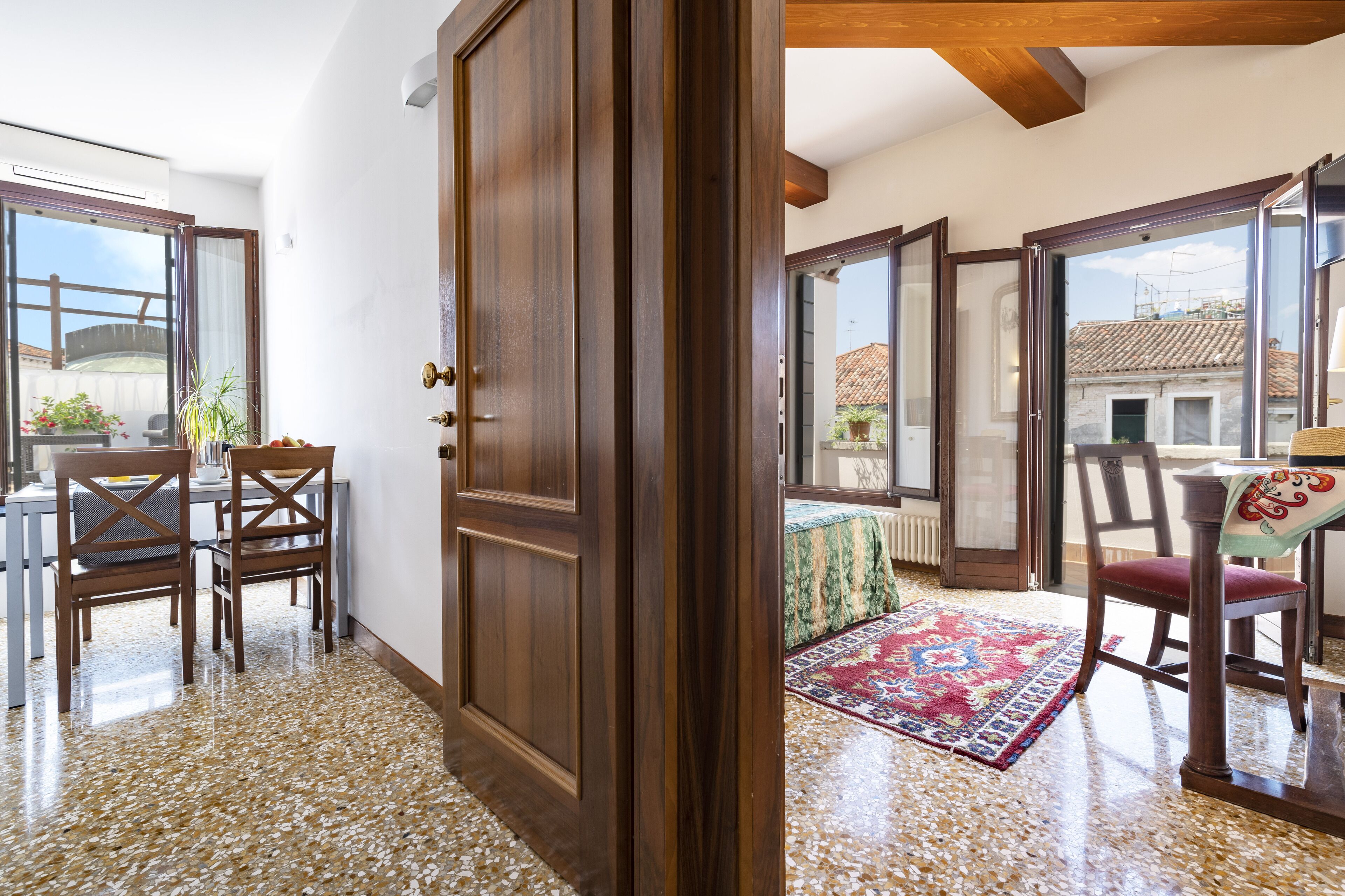 Une pièce avec une armoire en bois, une table à manger avec des chaises, un tapis à motifs et une vue sur un immeuble à travers une porte vitrée.