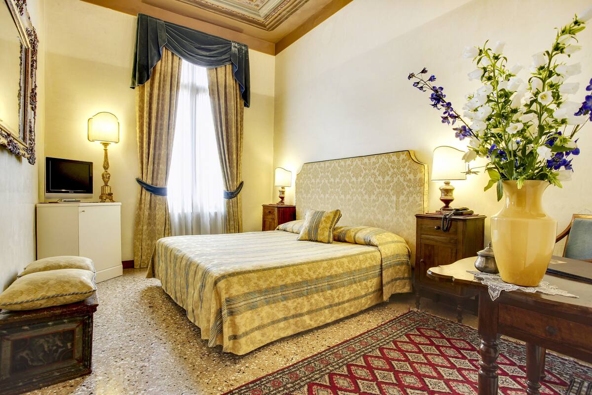 A hotel room with a large bed, a television, a desk with a lamp, and a vase with flowers.