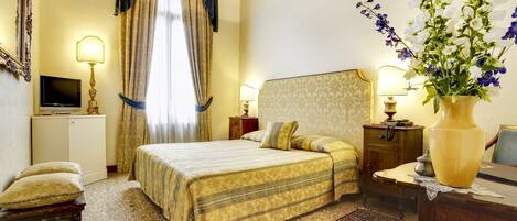 A hotel room with a large bed, a television, a desk with a lamp, and a vase with flowers.