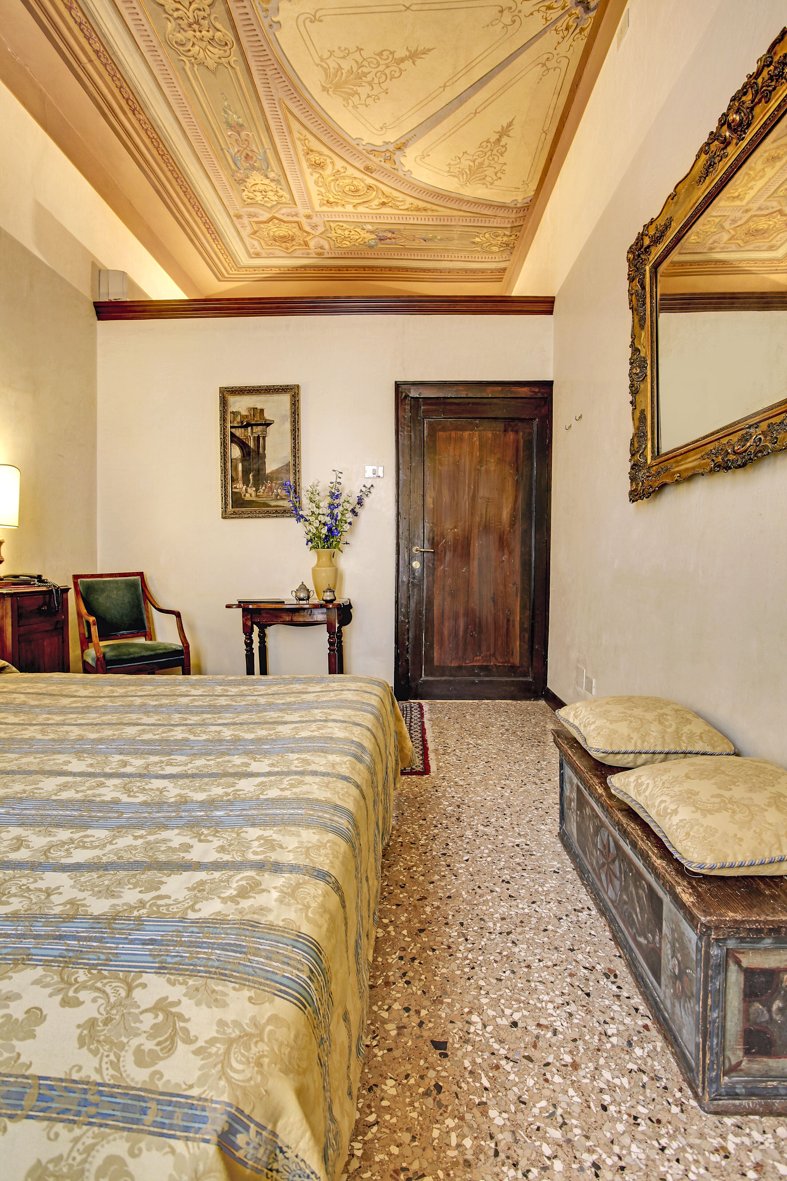 Ein Hotelzimmer mit einem großen Bett, einer Holztür, einem Spiegel, einem Sessel und einem kleinen Tisch mit einem Blumenstrauß.