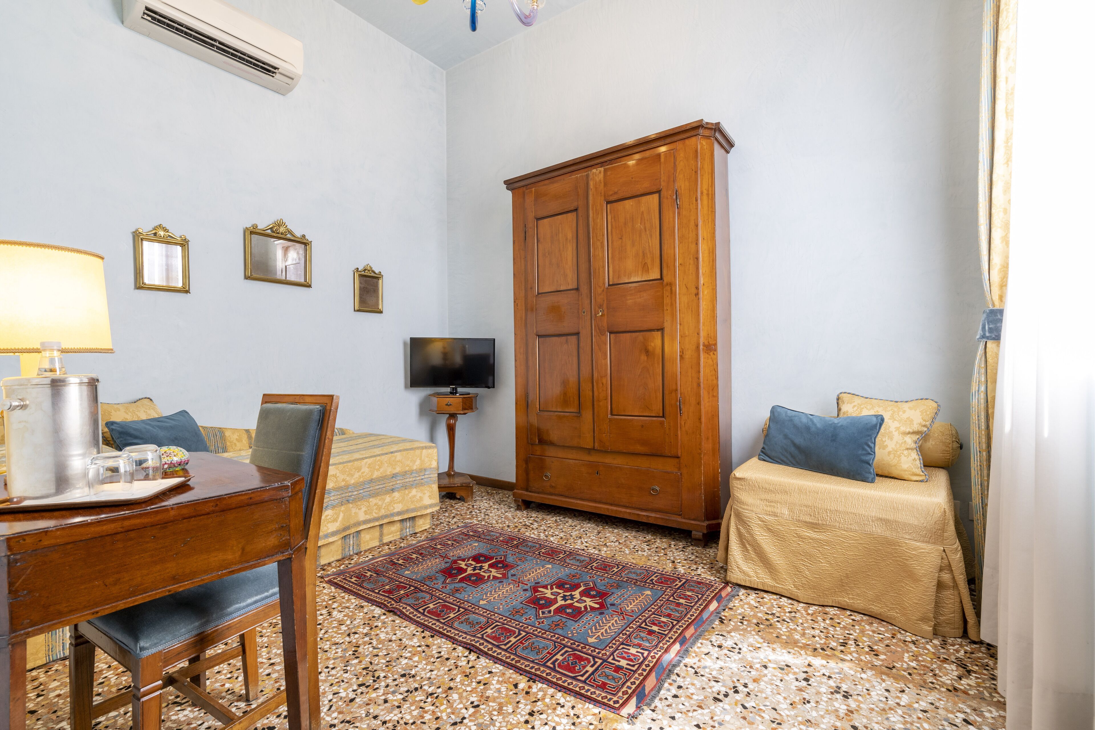 Una habitación con escritorio de madera, silla y televisor sobre un mueble bajo. Se observa un armario, una cama con colcha estampada y una alfombra decorativa.
