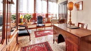 A spacious room with a large wooden desk, a comfortable armchair, and a chandelier. There are large windows with curtains, a red patterned rug, and a view of the outside.