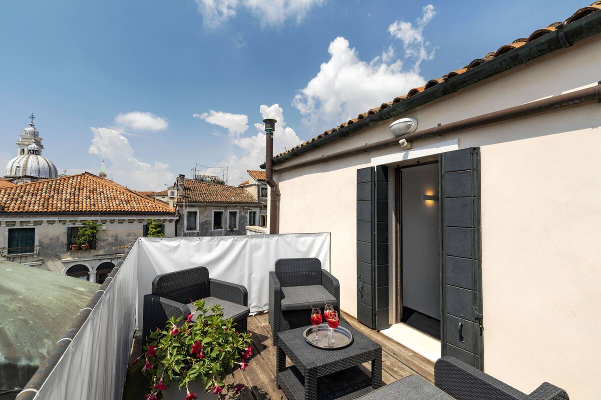 A rooftop terrace with wicker furniture, a table with glasses, and a view of historic buildings.