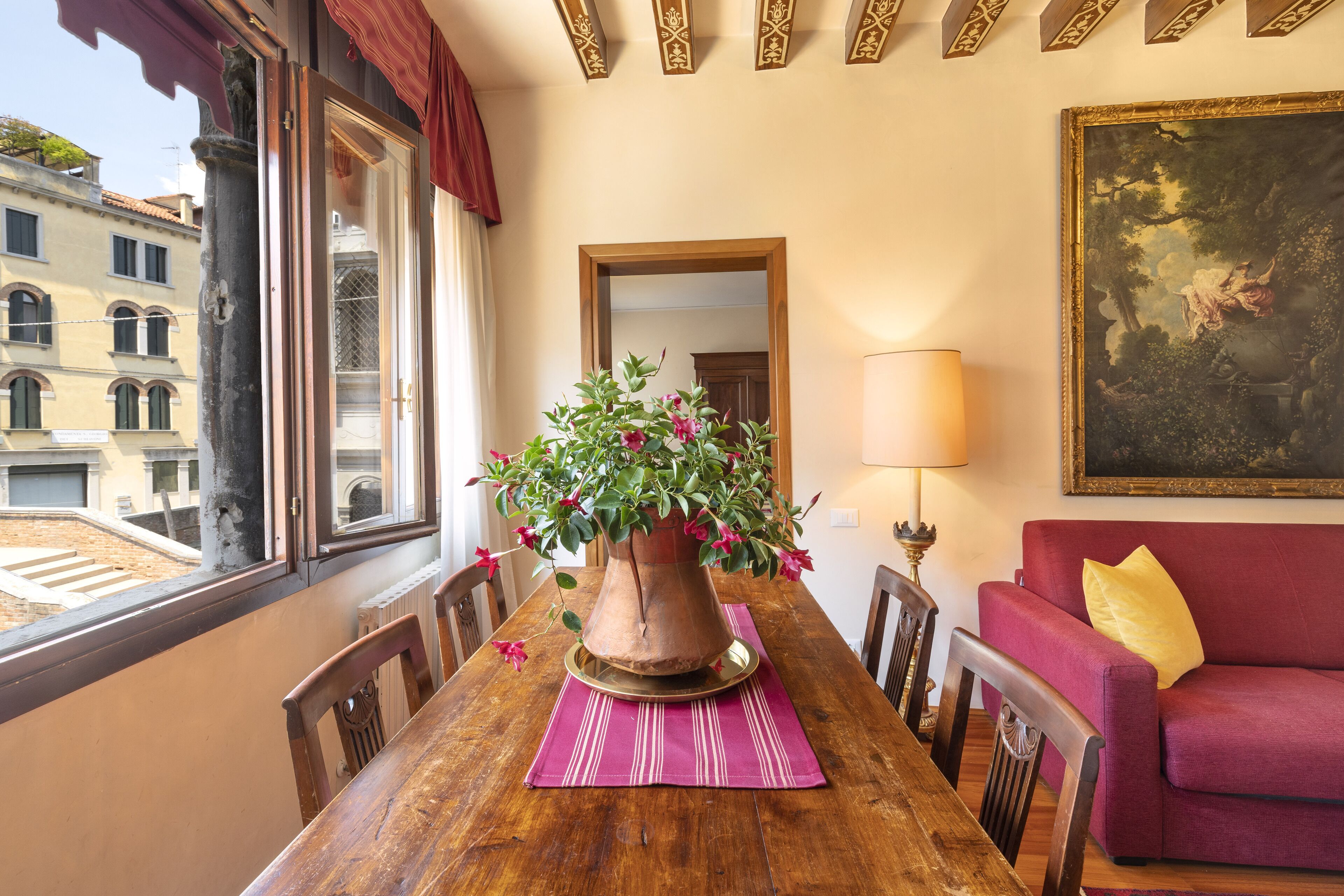 A dining table with a potted plant, a red sofa, and a framed painting on the wall.