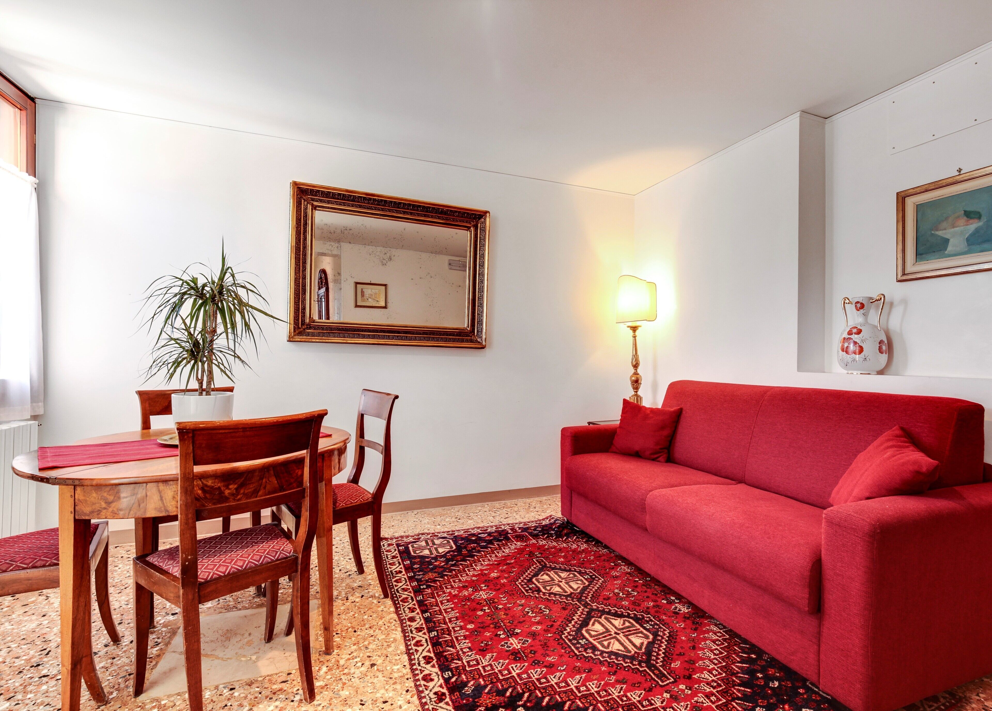 A living room with a red sofa, a wooden dining table with chairs, a potted plant, a mirror, and a lamp.
