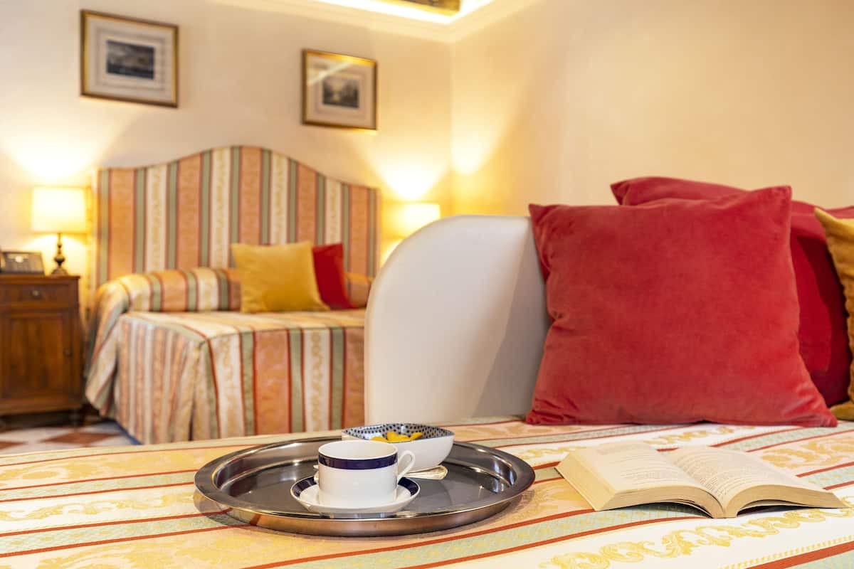 A hotel room with a bed, a bedside table with a lamp, a tray with a cup and saucer, and an open book on a patterned bedspread.