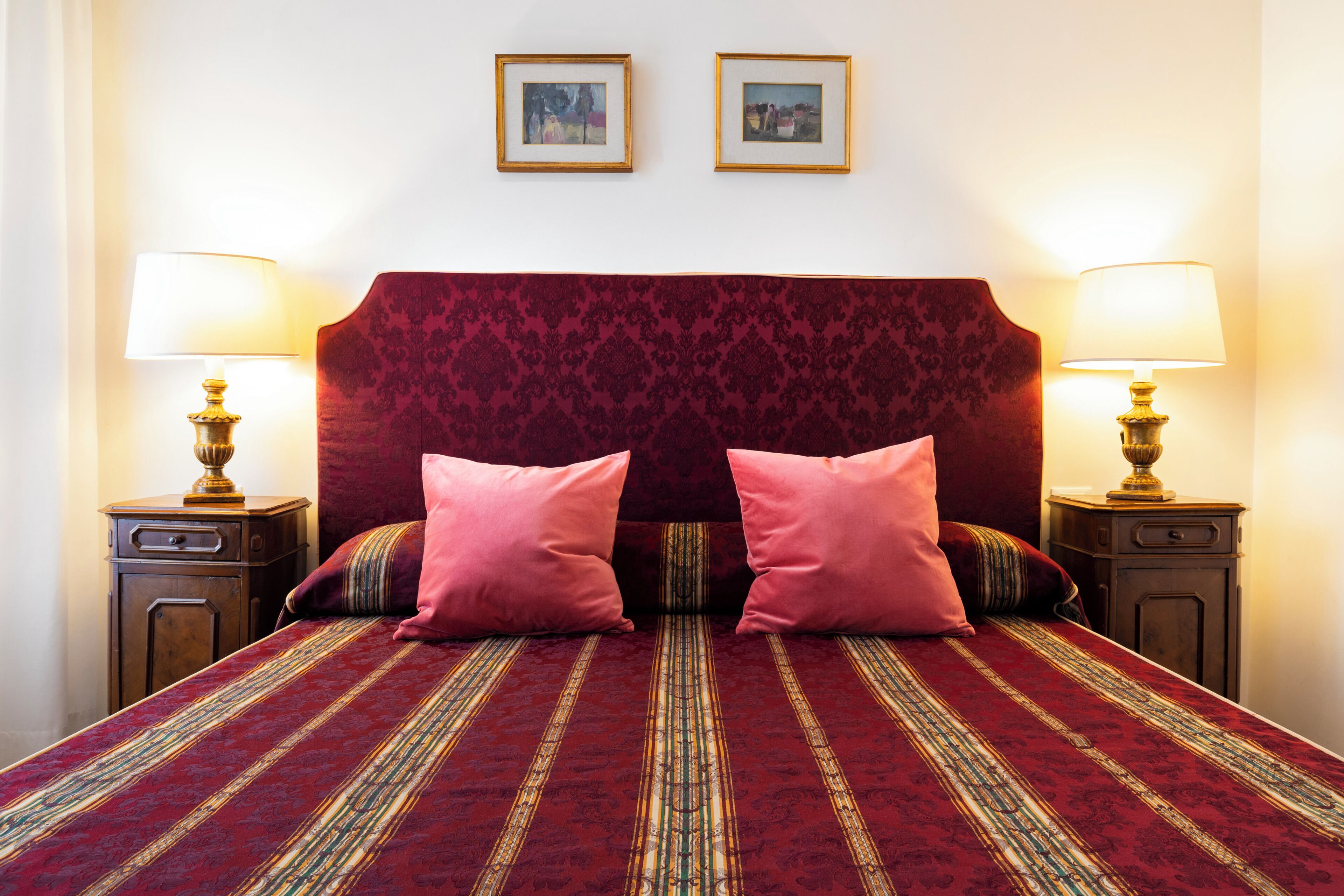 A double bed with a red and gold patterned bedspread, two pink pillows, and two nightstands with lamps.