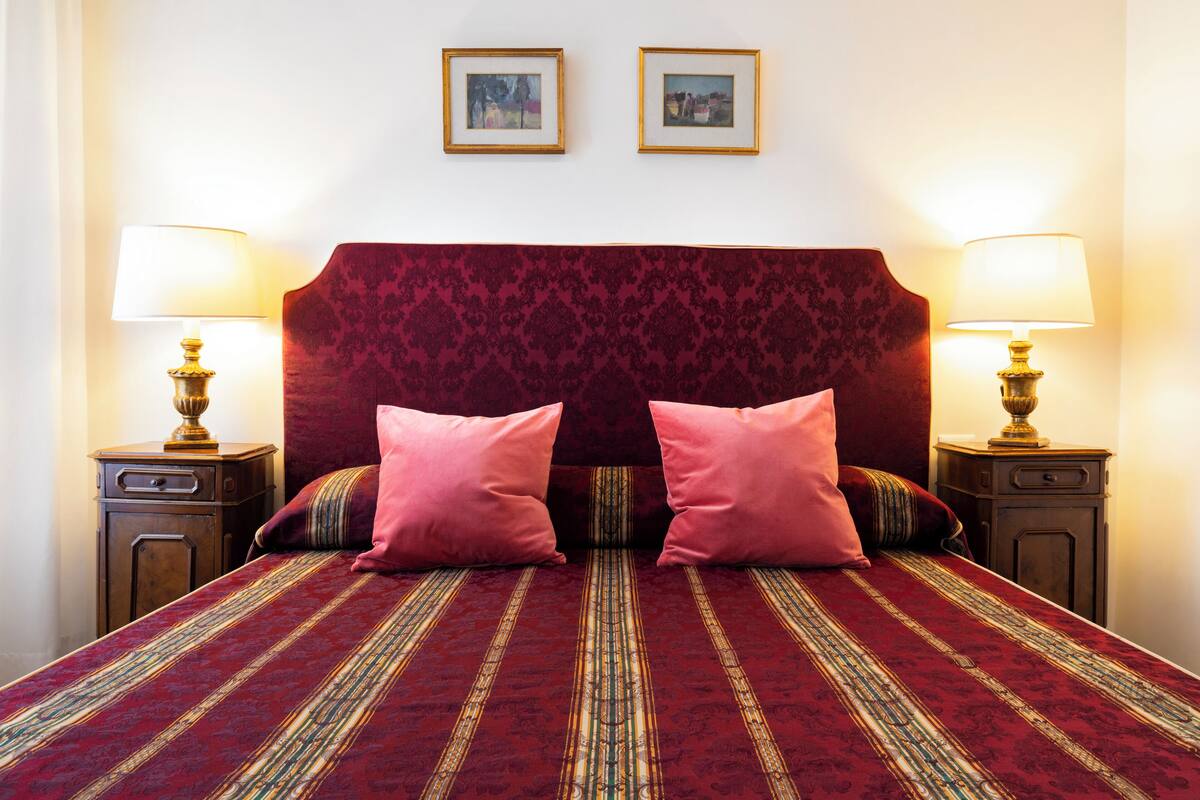 A double bed with a red and gold patterned bedspread, two pink pillows, and two nightstands with lamps.