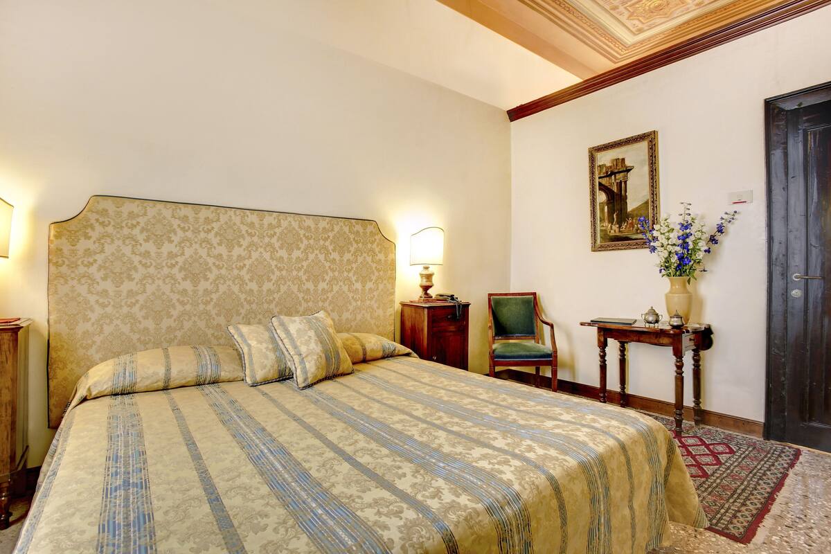 A bedroom with a large bed, a nightstand, a chair, a desk, a vase with flowers, and a framed picture on the wall.