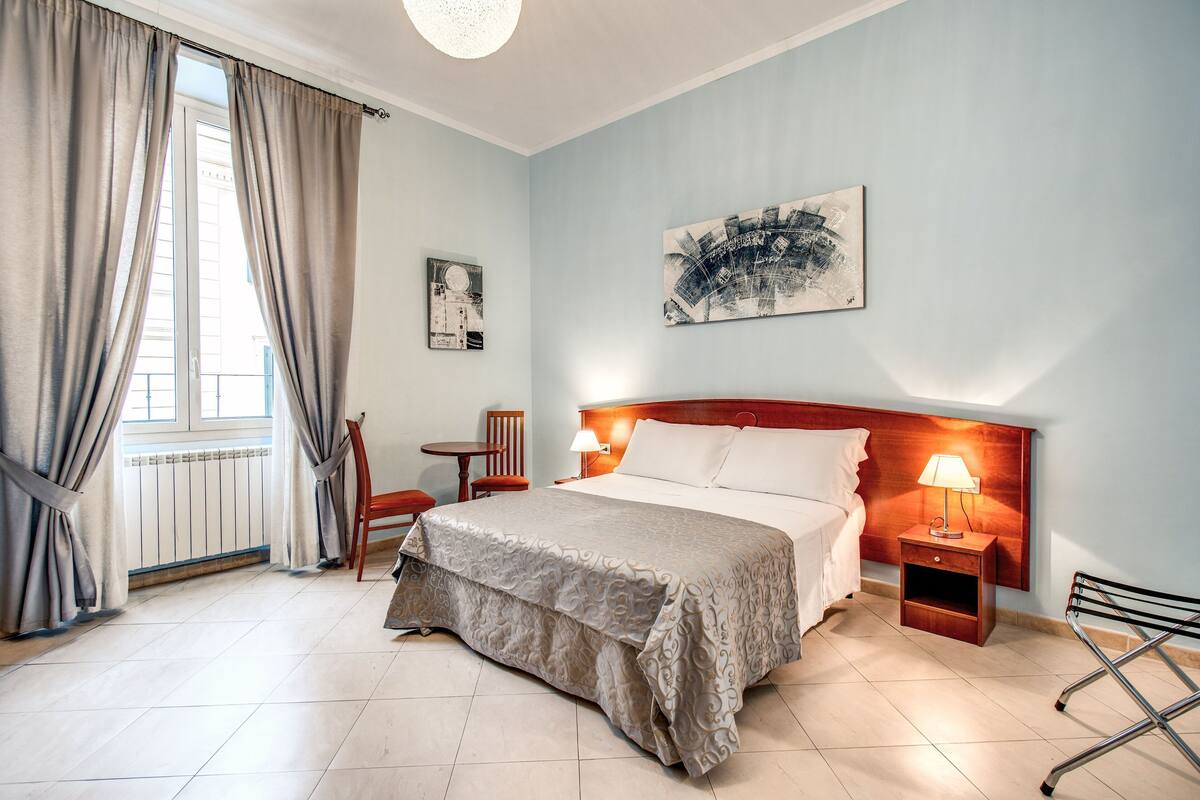 A hotel room with a bed, bedside tables, a chair, a window with curtains, and artwork on the wall.