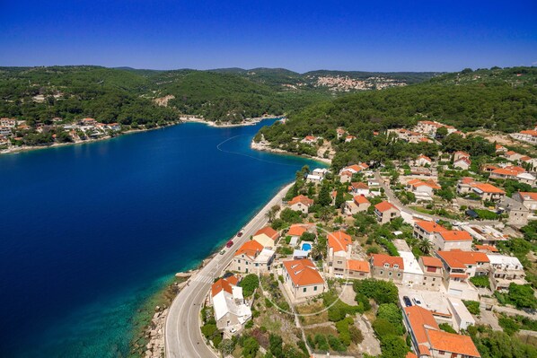 An aerial view of a coastal town with a clear blue sea, a winding road along the coast, and buildings with red-tiled roofs.
